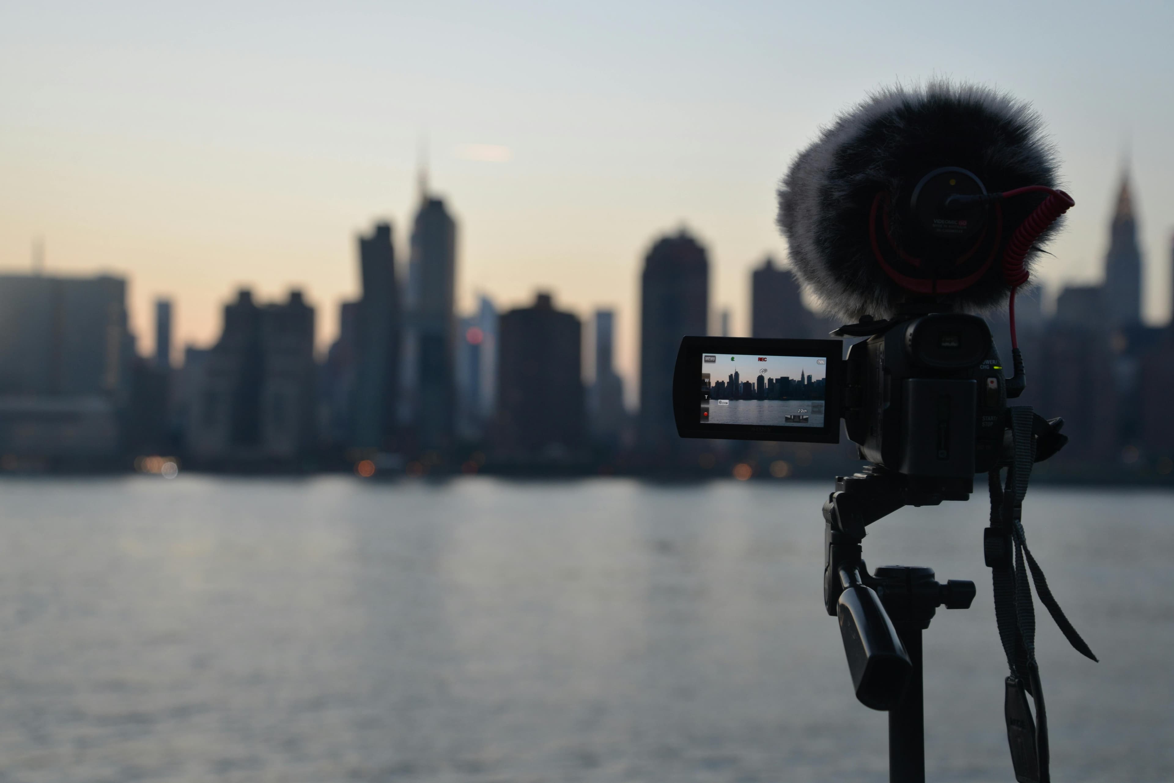 A video camera with a microphone on a tripod, recording a skyline at sunset.